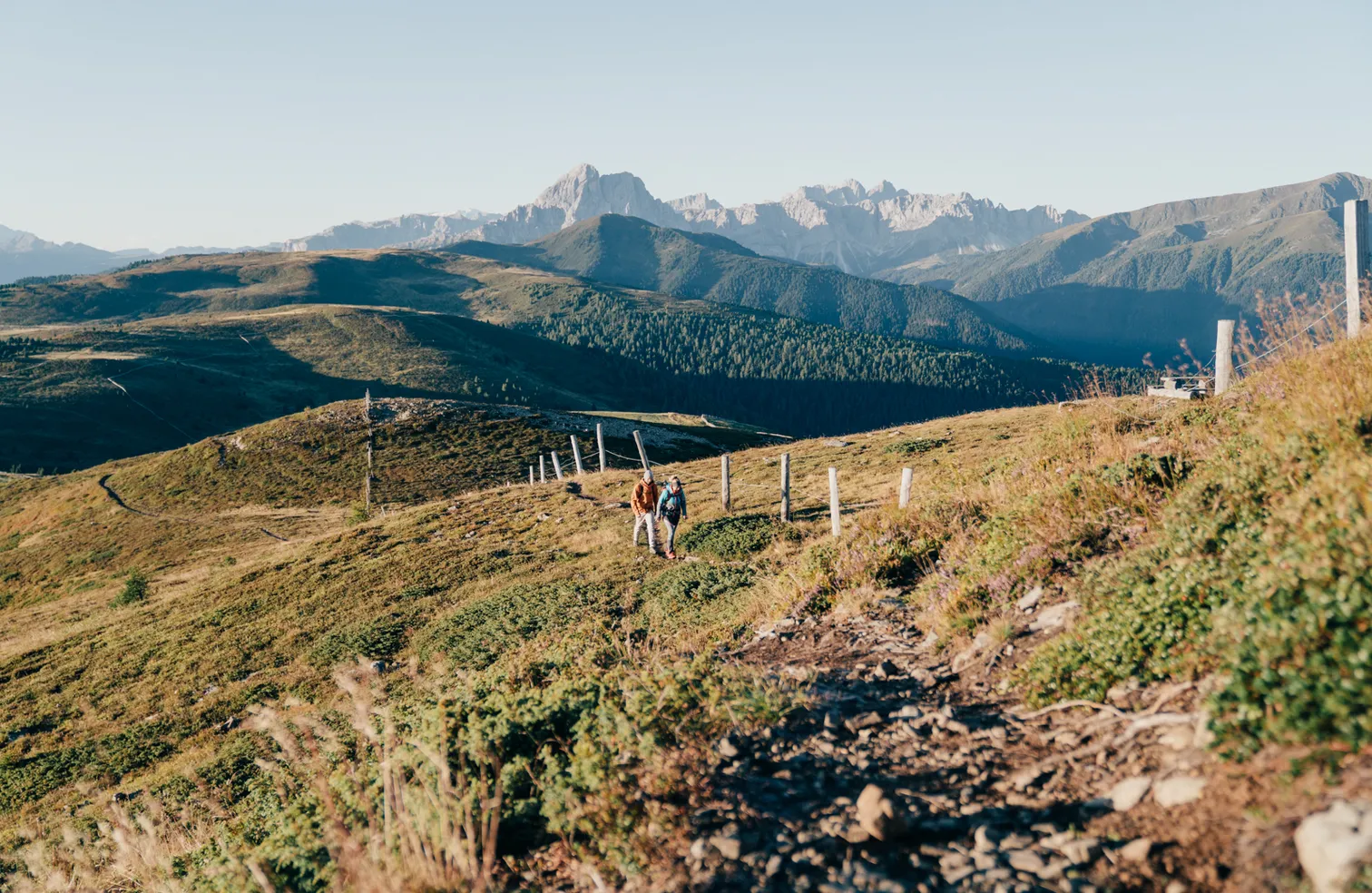 Two hikers on high trail above alpine meadows with panoramic view of the Geisler Dolomites in autumn near Luson
