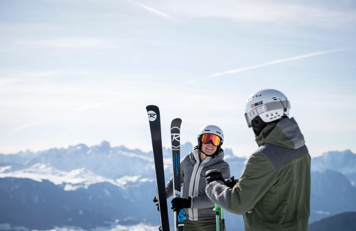 Smiling female skier with Rossignol skis and companion with Bliz helmet in front of snow-covered Dolomite panorama under clear sky in Villnöß