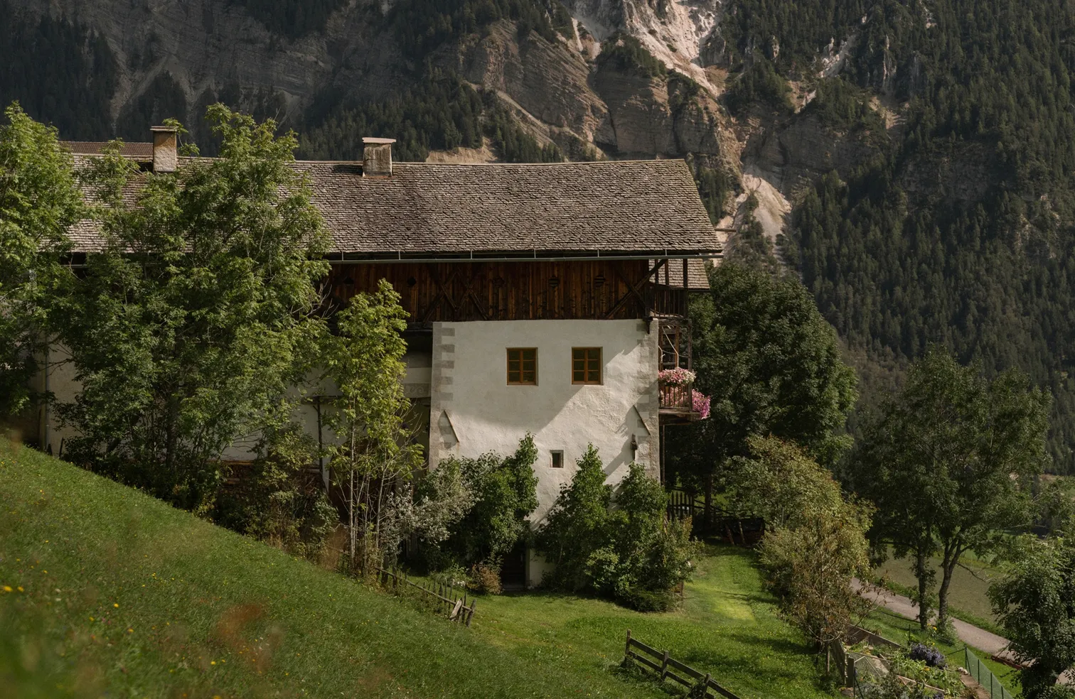 Tradizionale maso altoatesino con balcone di gerani davanti a fondale montano in estate, Funes-Luson