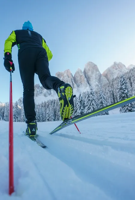 Langläufer im Skating-Schritt aus Froschperspektive auf Loipe in Villnöß mit Geislerspitzen und verschneitem Nadelwald