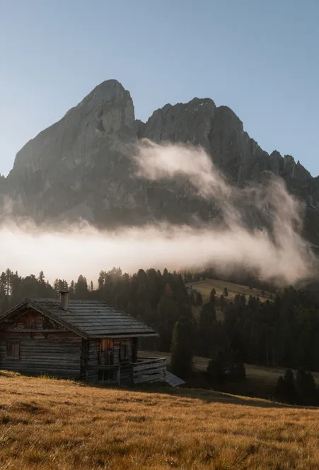 Wooden hut on alpine meadow in front of Peitlerkofel with dissolving morning fog in autumn