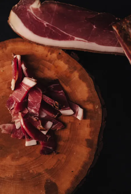 Südtiroler Speck aufgeschnitten auf einer runden Holzscheibe neben einem ganzen Speckstück vor dunklem Hintergrund
