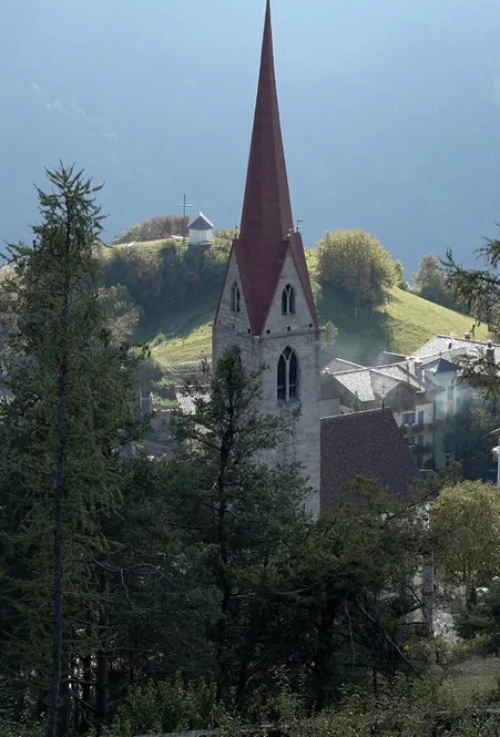 Gotischer Kirchturm mit rotem Spitzhelm in Villnöß, dahinter grüner Hügel mit Kapelle im morgendlichen Dunst der Dolomiten