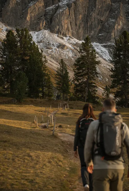 Two hikers on alpine trail towards Geisler rock faces in autumn