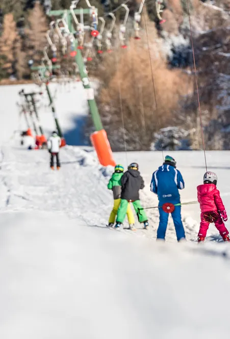 Children in colorful ski suits ride up on ski lift – ski school in sunshine with snow-covered forest in background