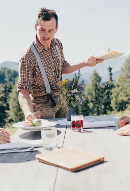 Almwirt in Lederhose serviert Südtiroler Gerichte an Wanderer am Holztisch mit Bergpanorama bei Villnöß