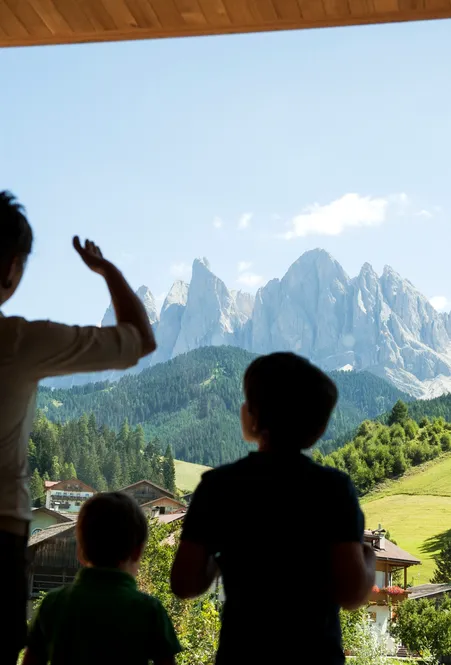 Famiglia con bambini guarda attraverso finestra panoramica verso Odle Dolomiti e prati verdi di Funes