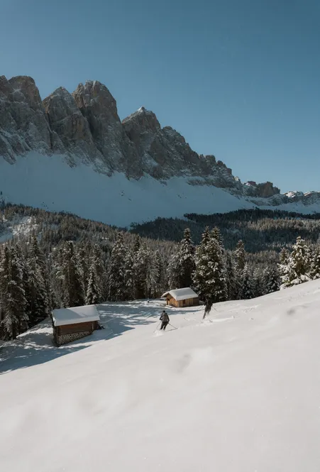 Scialpinista presso baite in legno su prato alpino innevato con vista sul gruppo delle Odle con cielo invernale limpido a Funes