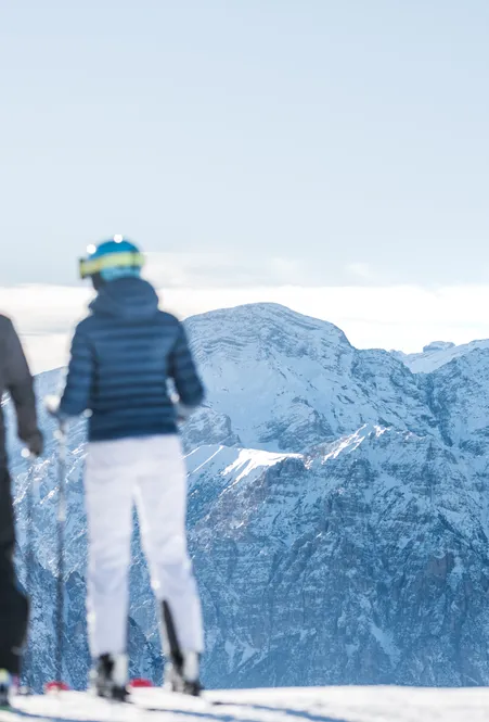Two skiers from behind look towards the snow-covered peaks of the Geisler Dolomites – cloud band over the valley in clear winter light