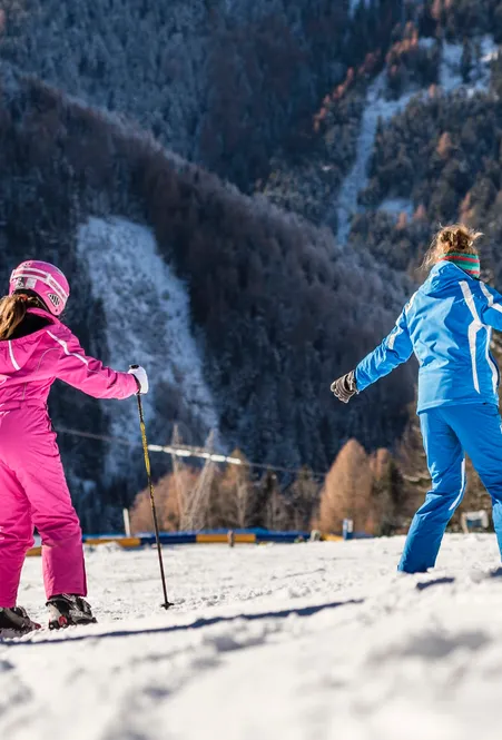 Ski instructor in blue suit shows girl in pink ski suit an exercise on flat slope in afternoon sun in Villnöß