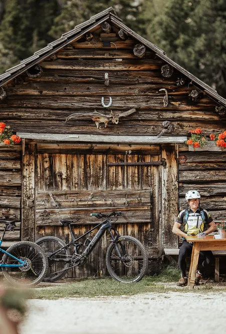 Mountainbiker bei Rast an rustikaler Holzhütte mit Hirschgeweih und Geranien im Villnößer Nadelwald
