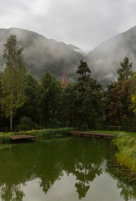 Natural bathing lake with wooden walkways and view of Luson bell tower in morning fog in front of South Tyrolean mountains