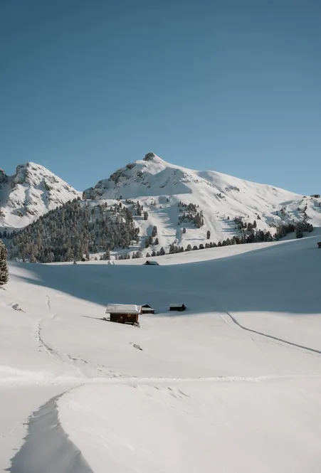 Paesaggio alpino profondamente innevato con baite in legno e tracce di scialpinismo davanti alle cime di Luson baciate dal sole con cielo azzurro