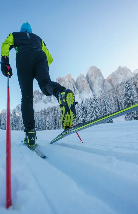 Fondista con tecnica skating dalla prospettiva della rana sulla pista a Funes con Odle e bosco di conifere innevato