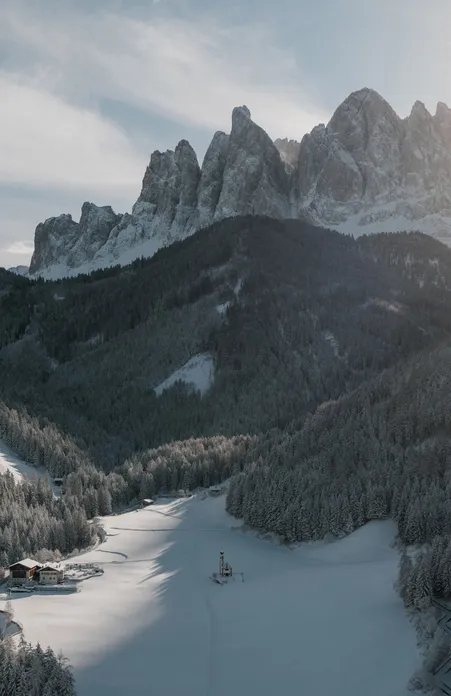 Winter Villnöß valley with snow-covered forests and the church of St. Magdalena in front of the Geisler Dolomites in sunlight