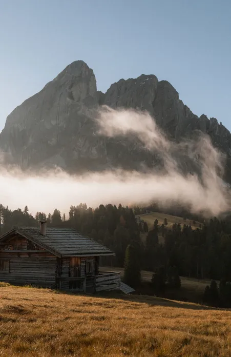 Baita in legno sul prato alpino davanti al Sass de Pütia con nebbia mattutina che si dissolve in autunno