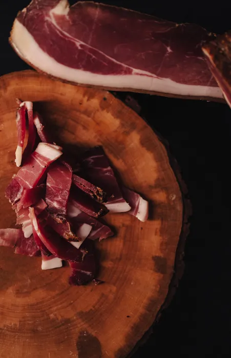 South Tyrolean speck sliced on round wooden board next to a whole piece of speck on dark background