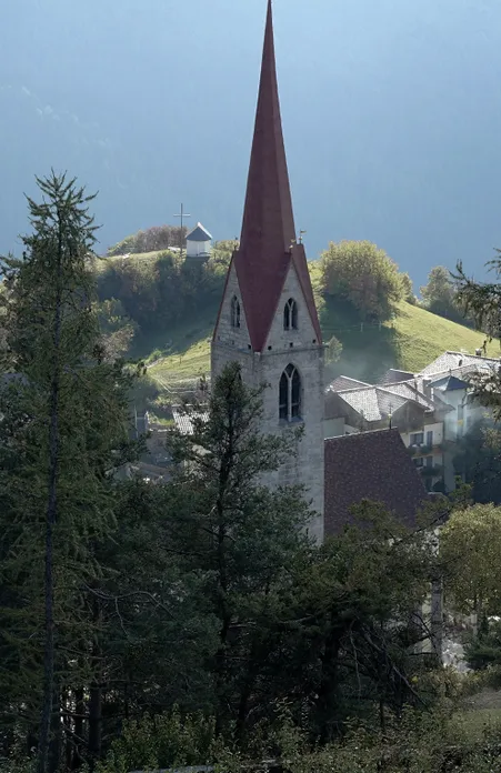 Gotischer Kirchturm mit rotem Spitzhelm in Villnöß, dahinter grüner Hügel mit Kapelle im morgendlichen Dunst der Dolomiten