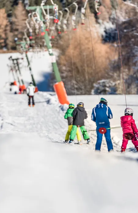 Bambini in tute da sci colorate salgono con lo skilift – scuola di sci al sole con bosco innevato sullo sfondo