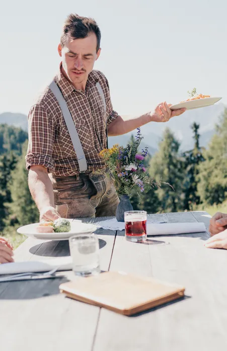 Alpine hut host in leather pants serves South Tyrolean dishes to hikers at wooden table with mountain panorama near Villnöß