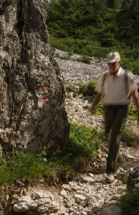 Hiker with cap walks along marked rock path over scree in the Geisler group of the Dolomites in summer