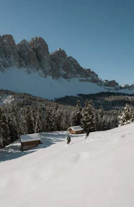 Ski tourer at wooden huts on snow-covered alpine meadow with view of Geisler group under clear winter sky in Villnöß