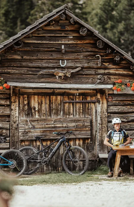 Mountain biker taking a break at rustic wooden hut with deer antlers and geraniums in Villnöß coniferous forest
