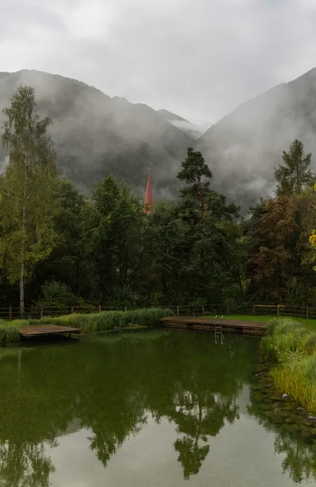 Laghetto naturale balneabile con passerelle in legno e vista sul campanile di Luson nella nebbia mattutina davanti ai monti dell'Alto Adige
