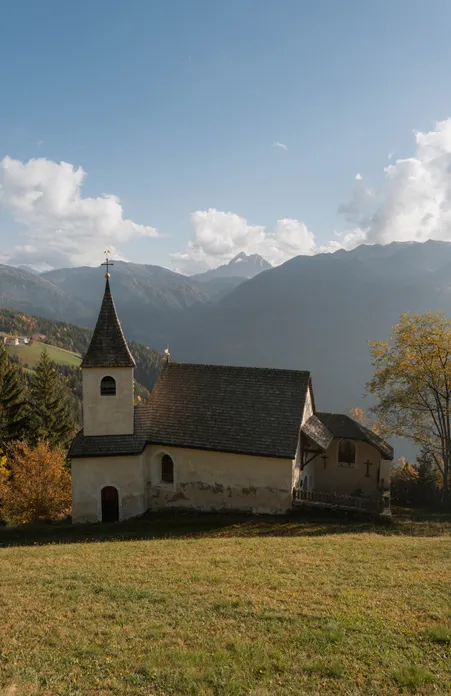 Cappella storica con tetto di scandole e campanile su prato autunnale con vista sulle catene montuose presso Luson in Alto Adige